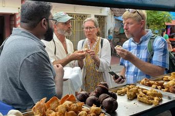 Three visitors standing at a street food stall eating fritters, with trays of pakoras and dark fried snacks on the counter in front of them and the vendor facing them.