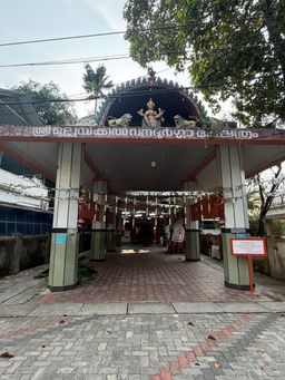 A covered entrance to a building with a tiled floor and pillars, featuring a decorative archway with a central figure flanked by two animal statues, and a sign with text above the entrance.