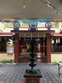 A temple entrance with a red-painted gateway topped by three colorful statues of deities. A tall black tiered oil lamp stands on a pedestal in the foreground on a tiled courtyard. Metal gates stand open behind the lamp, leading into the shrine area. A notice board is mounted on the left pillar, and a hanging light fixture is visible from the ceiling above.