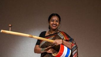 A woman in a dark sari laughing as she plays a long stringed folk instrument with a red, white and blue drum resting against her hip.