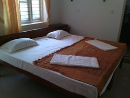 A bedroom with a double bed dressed in white sheets and an orange patterned throw, two folded white towels on the bed, a barred window with yellow curtains, and a small side table.