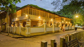 Two-storey cream and yellow colonial-style building at dusk, its windows lit, behind a white boundary wall with decorative pillars beside a paved street.