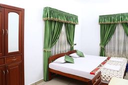 Bedroom with a wooden double bed under white sheets and green cushions, green curtains with gold trim, a tall wooden wardrobe with a mirror and a patterned bedspread at the foot.
