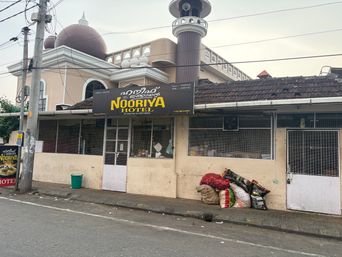 A single-story building with a signboard above the entrance stands beside a paved road. The building has a domed roof structure and a tall tower-like feature behind it. The front facade includes windows covered with wire mesh, a central doorway, and a white door on the right side. Several large sacks and bags are piled on the sidewalk near the right side of the building, next to a green bin on the left. Overhead electrical wires run across the sky above the scene.