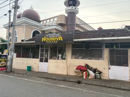 A single-story building with a signboard above the entrance stands beside a paved road. The building has a domed roof structure and a tall tower-like feature behind it. The front facade includes windows covered with wire mesh, a central doorway, and a white door on the right side. Several large sacks and bags are piled on the sidewalk near the right side of the building, next to a green bin on the left. Overhead electrical wires run across the sky above the scene.