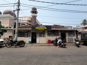 A street-level view of a single-story building with a sign above the entrance, set beneath overhead power lines. To the left, a larger structure with domes and a tall minaret rises behind the building. Several motorcycles are parked along the roadside in front, and a person sits on one of them near the entrance. A red sack rests on the ground beside the doorway, and a utility pole stands on the left side of the frame.