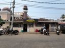 A street-level view of a single-story building with a sign above the entrance, set beneath overhead power lines. To the left, a larger structure with domes and a tall minaret rises behind the building. Several motorcycles are parked along the roadside in front, and a person sits on one of them near the entrance. A red sack rests on the ground beside the doorway, and a utility pole stands on the left side of the frame.