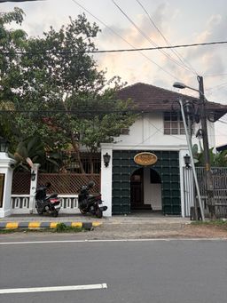 A white two-story building with a tiled roof stands behind a green double gate with an arched opening. Two scooters are parked on the sidewalk in front of the gate, next to a low fence with a lattice panel. A streetlight pole stands to the right of the gate, and overhead wires cross the sky above the building. Trees with green leaves grow behind the fence, and a yellow and black striped curb runs along the edge of the road in the foreground.