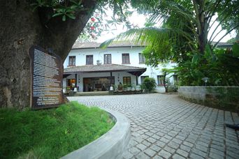 A white two-storey colonial building with a tiled-roof porch, set across a cobbled forecourt and framed by a large tree and palms, with a bronze plaque on a wooden board to the left.