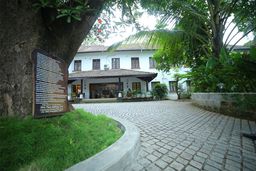 A white two-storey colonial building with a tiled-roof porch, set across a cobbled forecourt and framed by a large tree and palms, with a bronze plaque on a wooden board to the left.