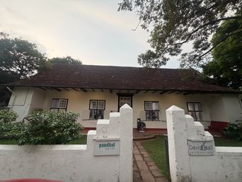 A single-story white building with a dark tiled roof and multiple windows with black frames stands behind a white wall with two gate pillars. A paved path leads from the foreground through an opening between the pillars toward the building’s entrance. Green bushes grow beside the wall on the left, and trees frame the scene on both sides under an overcast sky. Small signs are mounted on each gate pillar.