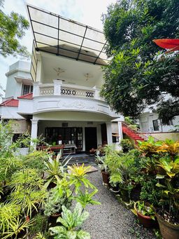 Two-storey white house with a curved balcony and a metal canopy, seen down a gravel path lined with potted plants and shrubs, with a red staircase at the side.