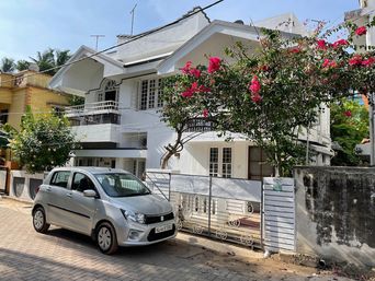 White two-storey house behind a pink-flowering tree and a decorative iron gate, with a silver hatchback parked on the paved street in front and a concrete boundary wall.
