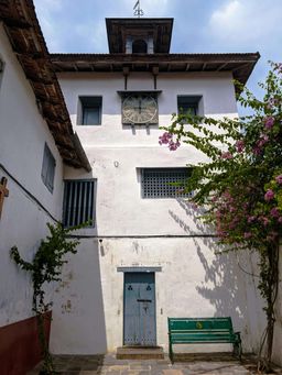 The synagogue’s white clock tower with a round clock face and weathervane, a bougainvillea and green bench below it.