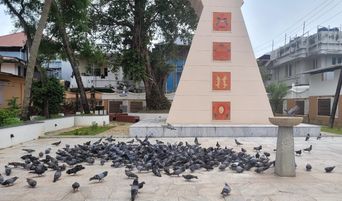 A large flock of pigeons on a paved courtyard in front of a tall painted pillar with panels of religious symbols.