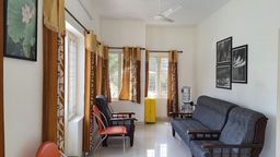 Sitting area with two grey-upholstered wooden sofas, an orange chair, gold patterned curtains, and a water dispenser beside framed black-and-white prints.