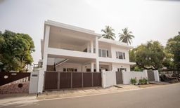 A white two-storey villa behind a dark wooden gate, with a covered upper balcony, a side staircase, and palm trees and large shade trees beside the road.