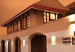White two-storey building at dusk with arched doorways, a red-tiled roof, dark-framed windows and wall lights along the facade.