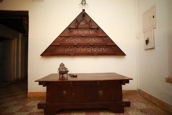 Heavy dark wooden chest with brass fittings and a small brass lamp on top, below a triangular wooden panel with carved lattice on a whitewashed wall. Patterned tiled floor.