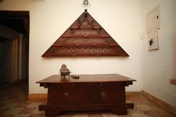 Heavy dark wooden chest with brass fittings and a small brass lamp on top, below a triangular wooden panel with carved lattice on a whitewashed wall. Patterned tiled floor.