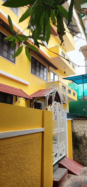 Yellow two-storey building with red-tiled awnings and dark-framed windows, behind a yellow boundary wall and a white ornate iron gate with steps up to it.