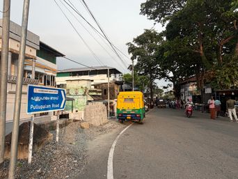A blue directional sign stands on the left side of a paved road, pointing toward a destination. A yellow and green auto-rickshaw drives away from the camera in the center of the road. On the right, several people stand near roadside shops under large trees, while others ride motorcycles further down the street. Buildings line both sides of the road, and overhead wires stretch across the sky.