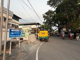 A blue directional sign stands on the left side of a paved road, pointing toward a destination. A yellow and green auto-rickshaw drives away from the camera in the center of the road. On the right, several people stand near roadside shops under large trees, while others ride motorcycles further down the street. Buildings line both sides of the road, and overhead wires stretch across the sky.