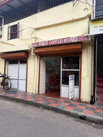 A street-level view of a coffee shop with a red sign above the entrance and a brown roll-down shutter partially raised. The shop front has glass doors and windows, one of which displays a poster with the shop’s name and a coffee cup graphic. Inside, a red chair and a small table are visible. To the left of the entrance, a white door with a metal grille is closed, and a bicycle is parked on the sidewalk beside it. The sidewalk is paved with red and grey tiles in a checkerboard pattern. A yellow sign is mounted on the wall to the right of the shop entrance, and overhead electrical wires run across the upper part of the building.