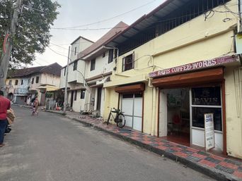 A street scene shows a row of buildings along a paved road. On the right, a yellow building has a shopfront with a sign above the entrance and a glass door with a poster inside. A bicycle is parked on the tiled sidewalk in front of the shop. Further down the street, other buildings line the road, and a person in a red shirt walks on the left side of the street while another person walks farther away. Trees and overhead wires are visible above the buildings under an overcast sky.