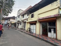 A street scene shows a row of buildings along a paved road. On the right, a yellow building has a shopfront with a sign above the entrance and a glass door with a poster inside. A bicycle is parked on the tiled sidewalk in front of the shop. Further down the street, other buildings line the road, and a person in a red shirt walks on the left side of the street while another person walks farther away. Trees and overhead wires are visible above the buildings under an overcast sky.