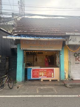 A small roadside shop with a tiled roof and a blue corrugated awning stands on a paved street. A person in a pink shirt stands inside the open front counter area. A large red sign with images of drinks is placed on the counter. A bicycle is parked on the left side of the shop. Overhead, numerous electrical wires crisscross against a bright sky. To the right, a closed metal shutter is visible, decorated with a garland of yellow and orange flowers.