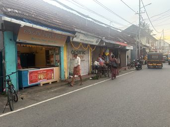 A street scene shows a row of small shops with tiled roofs along a paved road. A person in a light shirt and a patterned lower garment walks past a shop with a closed metal shutter decorated with a garland. Another person stands near a bicycle loaded with goods. A third person in a dark outfit walks away down the road. A bicycle is parked on the left side near a shop counter with a red advertisement panel. Further down the street, a three-wheeled vehicle and other vehicles are visible, with overhead wires crisscrossing above.