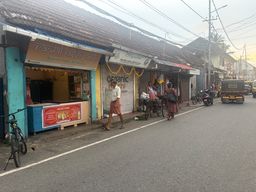 A street scene shows a row of small shops with tiled roofs along a paved road. A person in a light shirt and a patterned lower garment walks past a shop with a closed metal shutter decorated with a garland. Another person stands near a bicycle loaded with goods. A third person in a dark outfit walks away down the road. A bicycle is parked on the left side near a shop counter with a red advertisement panel. Further down the street, a three-wheeled vehicle and other vehicles are visible, with overhead wires crisscrossing above.