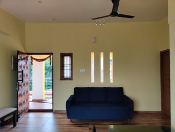 Living room with pale yellow walls, a navy three-seater sofa on a wood floor, a ceiling fan, and a carved wooden door open to a balcony.
