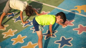 Two children bending forward with hands on the ground, playing on a painted playground surface covered in blue, yellow and pink stars.