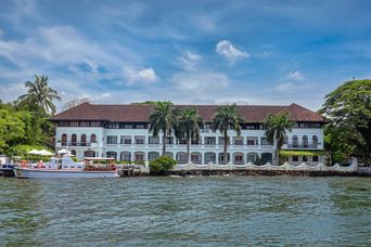 White colonial-style building with a red-tiled roof and arched windows, set at the water’s edge behind palm trees, with a white boat moored in front.