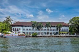 White colonial-style building with a red-tiled roof and arched windows, set at the water’s edge behind palm trees, with a white boat moored in front.