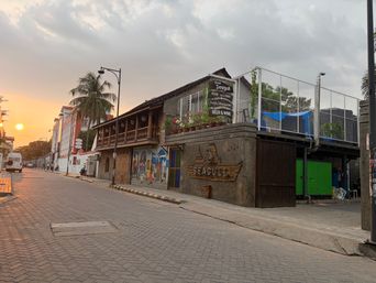 A two-story building with a wooden upper level and a stone lower level stands on a paved street at sunset. A sign on the upper level lists food and drink options, and a wooden carving on the lower level features a ship and a name. A mural with colorful figures is painted on the wall beside the entrance. A metal fence encloses a rooftop area with a blue tarp, and a green door is visible on the side. Palm trees line the street, and a vehicle is parked in the distance under a cloudy sky with the sun low on the horizon.