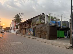 A two-story building with a wooden upper level and a stone lower level stands on a paved street at sunset. A sign on the upper level lists food and drink options, and a wooden carving on the lower level features a ship and a name. A mural with colorful figures is painted on the wall beside the entrance. A metal fence encloses a rooftop area with a blue tarp, and a green door is visible on the side. Palm trees line the street, and a vehicle is parked in the distance under a cloudy sky with the sun low on the horizon.