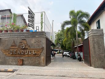 A concrete gateway with a carved wooden sign mounted on its left pillar opens onto a paved alleyway. To the left of the entrance, a raised wall topped with potted plants and a wire fence supports a vertical signpost. On the right, a matching concrete pillar stands beside an open gate, with parked motorcycles and a white car visible further down the lane. Tall palm trees and other greenery line the right side of the path, leading toward buildings in the distance under an overcast sky.