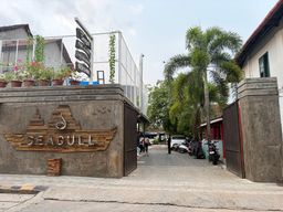 A concrete gateway with a carved wooden sign mounted on its left pillar opens onto a paved alleyway. To the left of the entrance, a raised wall topped with potted plants and a wire fence supports a vertical signpost. On the right, a matching concrete pillar stands beside an open gate, with parked motorcycles and a white car visible further down the lane. Tall palm trees and other greenery line the right side of the path, leading toward buildings in the distance under an overcast sky.