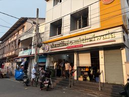 A street-level view of a two-story commercial building with a shop on the ground floor. The shop has a wide entrance with steps leading up to it, and several people are standing inside and near the doorway. To the right of the entrance is a closed metal roller shutter. Above the shop, the building has windows with bars and a signboard with lettering and a small emblem. On the left side of the street, a utility pole stands with wires running across the sky, and a few motorcycles are parked along the curb. A person on a motorcycle is positioned near the center of the frame, facing away from the camera. The adjacent building to the left has balconies and a covered walkway.