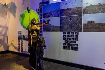 A costumed figure in dark ritual dress with a green backlit disc behind it, standing before a gallery wall covered in photographs of a landfill, at SMS Hall.