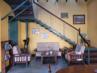 A cafe seating area with floral-upholstered sofa and armchairs below a blue open staircase, framed pictures on a yellow wall.