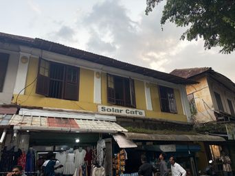A two-story yellow building with white trim stands under a cloudy sky, its upper floor featuring several windows with dark shutters. A sign is mounted on the facade below the windows. On the ground level, a shop with a corrugated metal awning displays clothing items hanging outside, while people stand and walk along the street in front. To the right, an adjacent building with a weathered exterior and a tiled roof is partially visible, along with tree branches extending into the upper right corner of the frame.