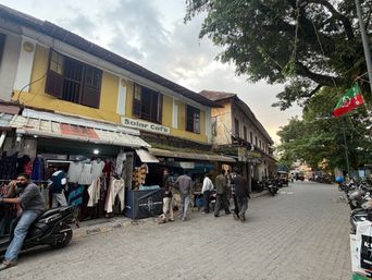 A street scene shows a row of two-story buildings with shops on the ground floor. On the left, a yellow building has a shop with clothes hanging outside and a sign above the entrance. Several people are standing near the shop entrance, and one person sits on a parked scooter in the foreground. Further down the street, more people walk along the paved road, and parked motorcycles line the right side. A flag hangs from a tree on the right, and the sky is partly cloudy.