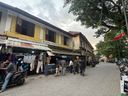 A street scene shows a row of two-story buildings with shops on the ground floor. On the left, a yellow building has a shop with clothes hanging outside and a sign above the entrance. Several people are standing near the shop entrance, and one person sits on a parked scooter in the foreground. Further down the street, more people walk along the paved road, and parked motorcycles line the right side. A flag hangs from a tree on the right, and the sky is partly cloudy.