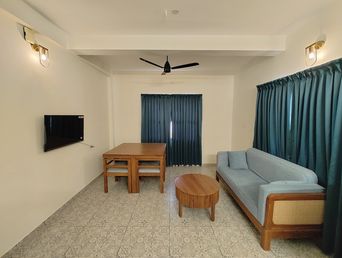 Living room with a wall-mounted TV, a small wooden dining table with two chairs, a round wooden coffee table, and a grey-blue sofa on a patterned tiled floor.