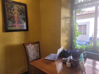 A corner table by a window at a cafe, with a laptop and coffee cups, a framed picture of Krishna on the yellow wall beside it.