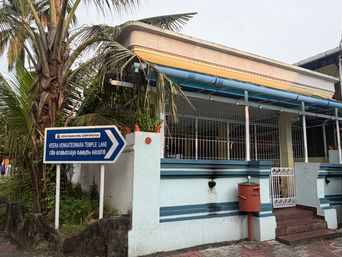A white building with blue trim stands behind a low wall topped with metal bars, with a blue and white directional sign on posts beside a palm tree, a red post box mounted on the wall, and steps leading up to a white gate.